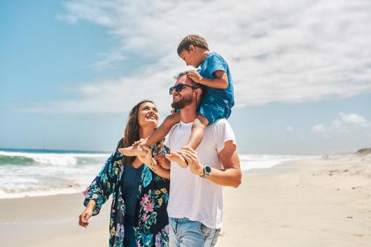 A smiling family walking along a summery beach with the father carrying his son on his shoulders