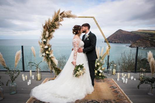 A bride and groom sharing a kiss surrounded by wedding decorations on the deck of the Venue at Sandy Cove hotel with a view over the bay behind them
